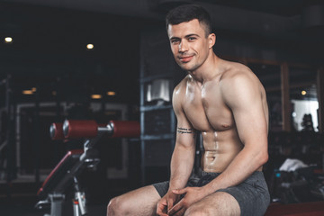 Fototapeta na wymiar Smiling tattooed sportsman is feeling exhausted after workout with machines indoors. He is sitting topless and relaxing. Copy space in left side