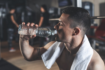 Fototapeta na wymiar Profile of sportsman quenching thirst from bottle. Tattooed shirtless man is using towel after workout. He is exercising with machines and heavy weights in gym