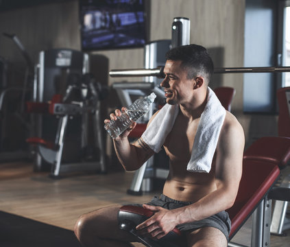 Merry sportsman is having break in hard training with barbell and machine. He is sitting on bench while having bottle with soft beverage. Healthy lifestyle with pleasure concept