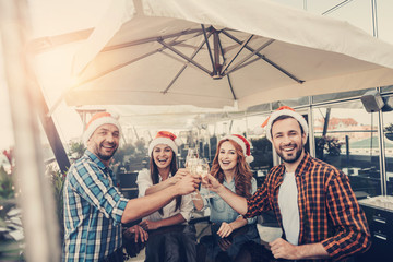 Best wishes. Two bearded men and lovely ladies in Santa hats sitting at the table and toasting. They holding glasses of champagne and smiling