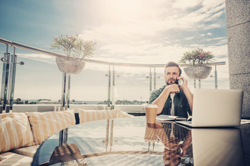 Important conversation. Handsome guy sitting at the table with laptop and having phone conversation. Beautiful sky, sunlight and balcony on background