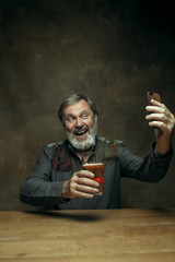 Smiling bearded man drinking alcohol in pub. Enjoying my favorite beer. The front view of handsome smiling senior man with glass of beer sitting at the wooden table. Studio shot with caucasian model