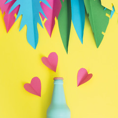 Painted in blue beer or cocktail bottle lies on a yellow background among hearts and palm tropical leaves made of paper.