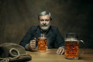 Smiling bearded man drinking alcohol in pub. Enjoying my favorite beer. The front view of handsome smiling senior man with glass of beer sitting at the wooden table. Studio shot with caucasian model