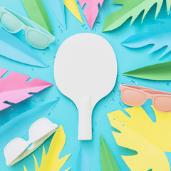 A tennis racket painted white lies among the tropical leaves of a palm tree made of paper and sunglasses. Summer concept of beach sports and recreation