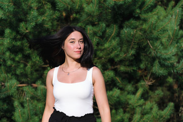girl in white shirt with black hair that develops in the wind against the background of coniferous trees