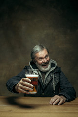 Smiling bearded man drinking alcohol in pub. Enjoying my favorite beer. The front view of handsome smiling senior man with glass of beer sitting at the wooden table. Studio shot with caucasian model