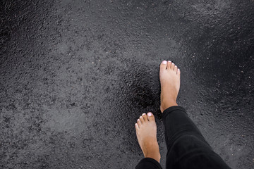 Young woman's barefoot walking on the wet, dark black asphalt after warm rain. Cloudy day in...
