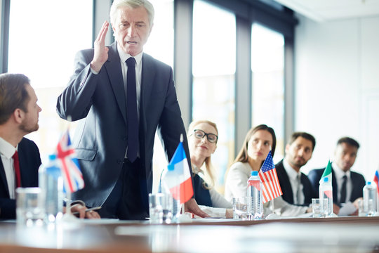 Aged confident politician in suit standing by table and debating with his foreign colleagues at political event