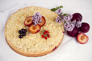 homemade pie with plums decorated with currants, strawberries and flowers on a white wooden table