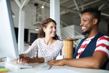 Smiling young businesswoman sitting at the workplace together with her partner and typing on...