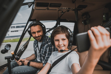 Portrait of happy bearded father and optimistic child taking photo on cellphone while wearing headsets. They spending time in helicopter
