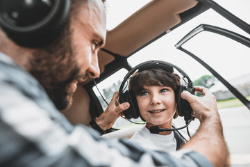 Satisfied bearded man putting modern headsets on cheerful boy while communicating with him. They sitting in modern helicopter inside