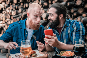 Curious men. Two bearded men feeling curious while reading news on the phone having tasty dinner
