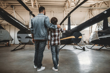 Full length kid pointing hand on big rotor plane. Man hugging him while listening his description...