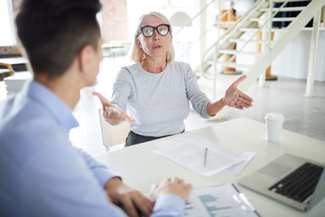 Obraz premium Mature female leader wearing eyeglasses sitting at desk and gesturing during a business meeting at office