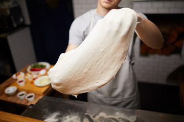 Young baker stretching dough while holding it over table during process of cooking pizza
