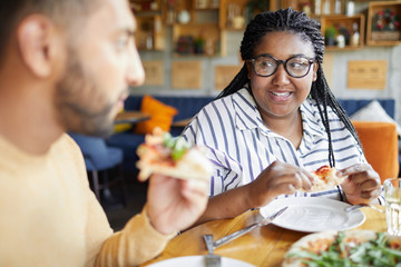 Two intercultural friends eating pizza by table and talking while sitting in cafe