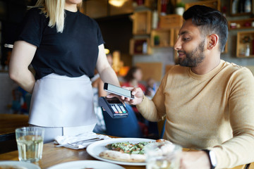 Young man holding his smartphone over payment machine while sitting by table in cafe during lunch