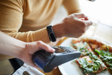 Hand of young man with smartwatch close to payment machine over served table during lunch