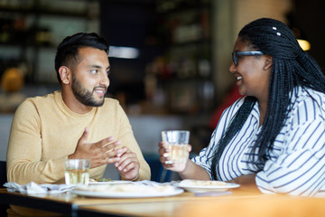 Intercultural guy and girl sitting by table in cafe, enjoying talk, drinks and lunch