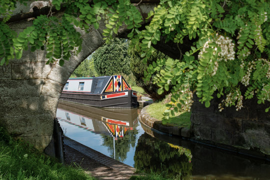 A close up view of the front part of a canal narrowboat. The view is taken through a old stone bridge and trees. The front of the leisure boat shows reflections in the water.