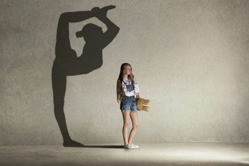 Baby girl dreaming about gymnast profession. Childhood and dream concept. Conceptual image with shadow of female gymnast on the studio wall