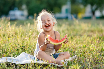 Little blond girl eating watermelon in the park.