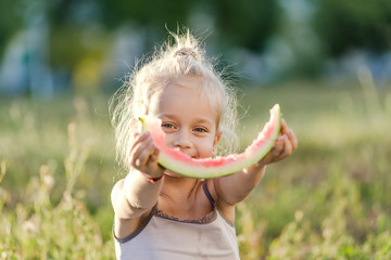 Little blond girl eating watermelon in the park.