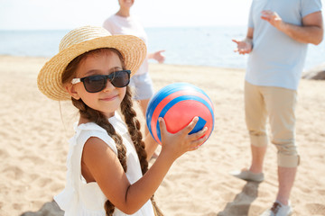 Adorable kid in hat and sunglasses holding ball while enjoying play with her parents by seaside