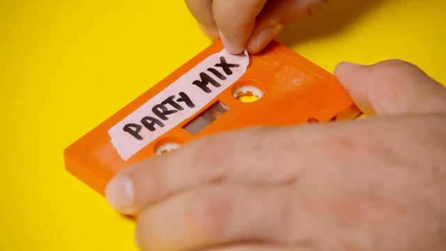 Applying a handwritten label on a vintage cassette tape (obsolete music technology) with the text Party Mix. Looks like magic but it's a simple reversed shot. Colorful scene.
