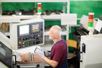 Mature engineer controlling process of work, he is using digital tablet while working on computer machine in factory