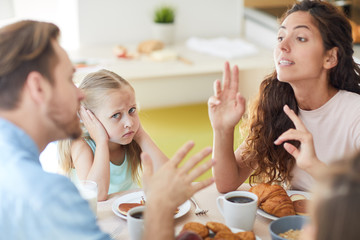 Young irritated wife saying something to her husband by breakfast during argument with little daughter between them