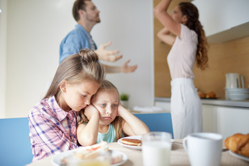 Two sad girls sitting by table in the kitchen while their parents having argument on background