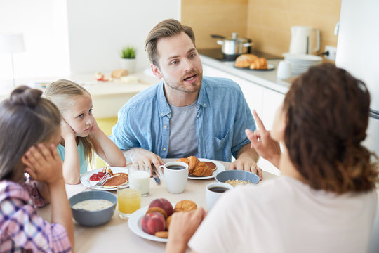 Young annoyed man rowing with his wife by breakfast in the kitchen with their two daughters near by