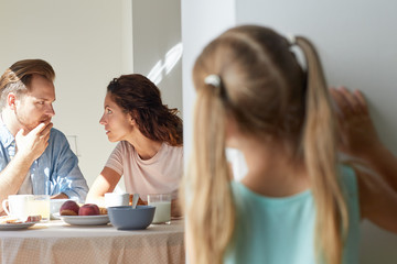 Young parents sharing secreta by breakfast while their little daughter standing by kitchen door and...