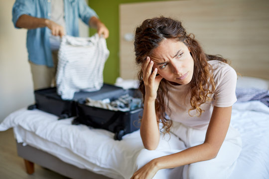Young scowling wife sitting on edge of bed on background of her husband preparing suitcase before leaving