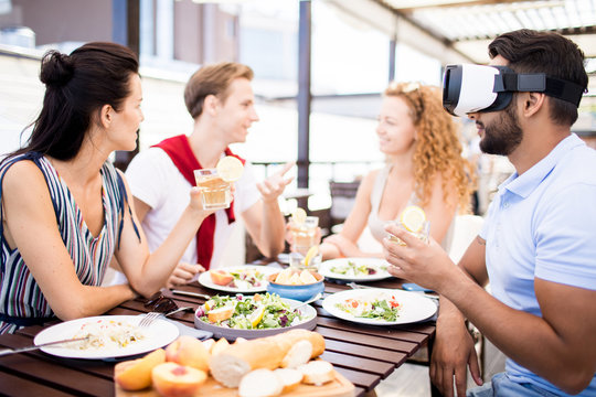 Young man in vr headset sitting by served table among his talking friends, watching video and having drink