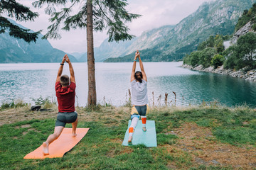 Fitness, sport, friendship and lifestyle concept - sportive couple making yoga exercises sitting on mats in cloudy morning outdoors near beautiful lake in camping, unusually nature, Norway.