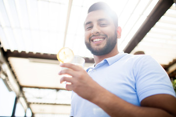 Young smiling man with glass of lemonade looking at you while enjoying sunny day in cafe