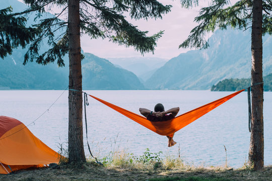 Back view of man silhouette relaxing on orange hammock between two trees pine enjoying the view at the lake in summer norwegian cloudy morning.