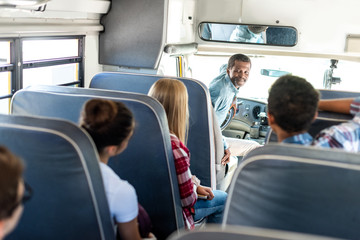 group of teen scholars riding school bus and talking with happy mature driver