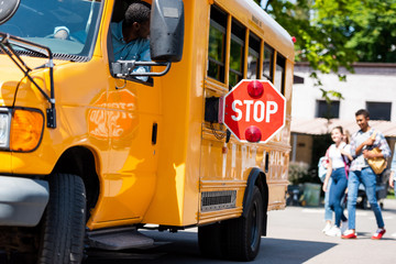 senior school bus driver looking at teens walking behind bus - obrazy, fototapety, plakaty