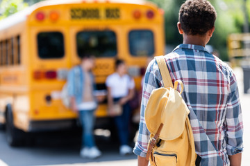 rear view of african american schoolboy walking to classmates leaning on school bus