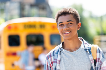 happy teen african american schoolboy looking at camera with blurred school bus on background