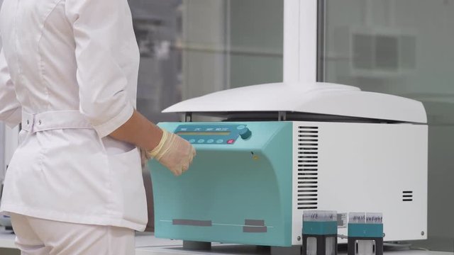 In the laboratory, a woman in a Bathrobe and gloves sets samples of human cells in test tubes in a centrifuge for unwinding and separating excess layers of cells