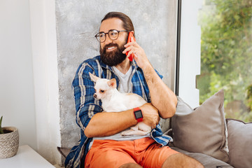 Calling wife. Dark-eyed smiling man feeling cheerful while calling his wife while holding little dog