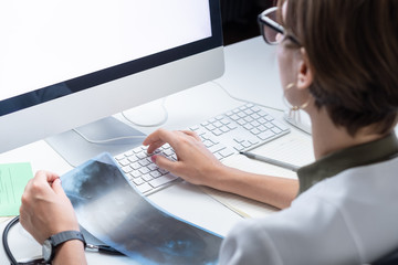 Digital health concept: young medical doctor working at a desktop computer. Female practitioner at modern medical doctor office examining x-ray and taking notes in computer.