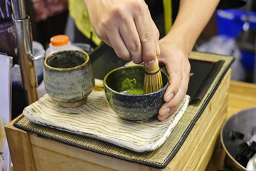 A man trying to mix a matcha green tea powder with hot water in a black bowl with a bamboo matcha whisk, demonstration of Japan green tea making art, Tools used for Japanese tea ceremony (chado).