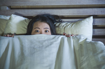 young scared and stressed Asian Chinese woman lying in bed suffering nightmare in fear and panic grasping blanket covering her horror face expression
